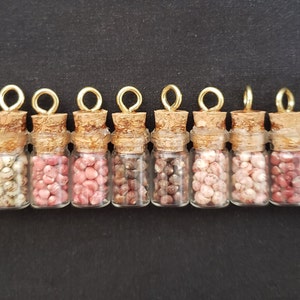 May include: Ten small glass bottles with cork tops and gold lids filled with various shades of pink and white beads.