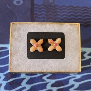 May include: A pair of flower-shaped earrings with a light peach color and a small pink center, presented in a gold-trimmed box with a black insert. The earrings are displayed on a white, fluffy material.