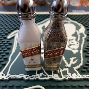 May include: Two small glass salt and pepper shakers shaped like bottles. The shakers are labelled "Johnnie Walker Red Label Blended Scotch Whisky". The shakers are sitting on a green and white bar mat.