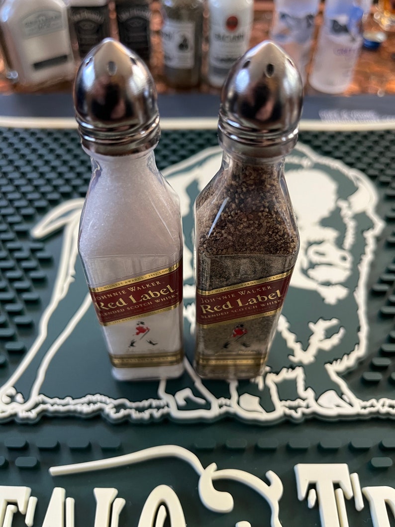 May include: Two small glass salt and pepper shakers shaped like bottles. The shakers are labelled "Johnnie Walker Red Label Blended Scotch Whisky". The shakers are sitting on a green and white bar mat.