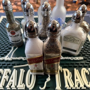May include: A collection of miniature liquor bottles repurposed as salt and pepper shakers. The bottles are filled with various spices and labelled with brand names such as Bacardi, Gentleman Jack, and Johnnie Walker Red Label.