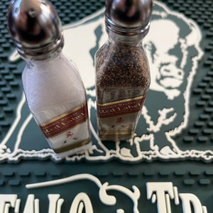 May include: Two small glass bottles with silver tops filled with salt and pepper. The bottles are labelled "Johnnie Walker Red Label" and are sitting on a green and white bar mat.