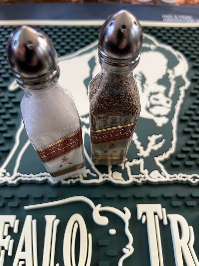 May include: Two small glass bottles with silver tops filled with salt and pepper. The bottles are labelled "Johnnie Walker Red Label" and are sitting on a green and white bar mat.