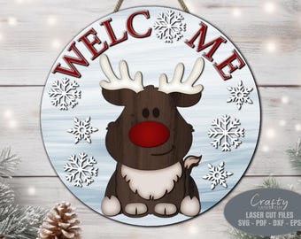 May include: A round wooden sign with a blue and white background. The sign says "Welcome" in red letters with a cartoon reindeer in the centre. The reindeer has a red nose and brown antlers. The sign is decorated with white snowflakes.