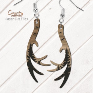 May include: A pair of wooden earrings shaped like antlers with a silhouette of pine trees and birds flying in the background. The earrings are brown with black accents.