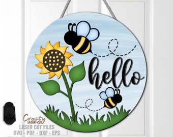 Op de afbeelding: Een rond houten bord met een blauwe en witte aquarel achtergrond. Een zonnebloem met een gele kern en zwarte zaden bevindt zich aan de linkerkant van het bord. De tekst "hello" is geschreven in zwarte cursieve letters. Twee cartoon bijen met zwarte lichamen, gele strepen en blauwe vleugels vliegen op het bord.