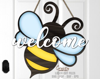 Op de afbeelding: Een houten bij deurhanger met blauwe vleugels en gele strepen. De bij heeft het woord "welcome" in wit op zijn lichaam.
