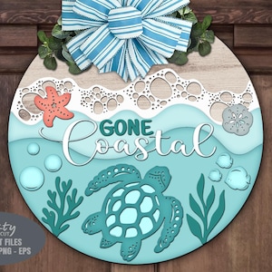 Op de afbeelding: Ronde houten bord met een kustthema. Het heeft een turquoise zee met een schildpad, bellen en zeewier. Het bord zegt "Gone Coastal" en is bekroond met een blauw-witte gestreepte strik en groen.
