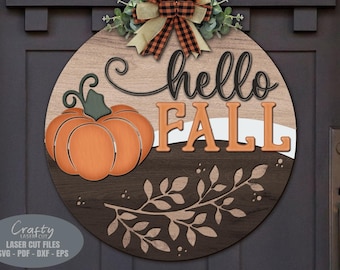 May include: A round wooden sign with the text "hello fall" in black and orange lettering. A carved pumpkin with a green stem is on the left side of the sign. A brown leafy branch is on the bottom of the sign.