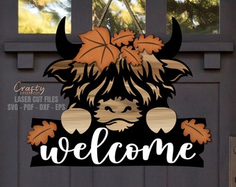 May include: A black and brown Highland cow with a welcome sign. The cow has a brown nose and is peeking over a wooden sign with the word "welcome" in white letters.