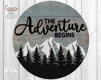 May include: A round wooden sign with a blue and grey woodgrain background. The sign features a black silhouette of mountains, trees, and the text "The Adventure Begins".