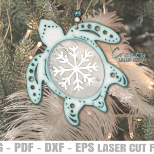 Può includere: Un ornamento a forma di tartaruga in legno tagliato al laser di colore blu-verde con un fiocco di neve bianco al suo interno. L'ornamento è appeso a un albero di Natale.