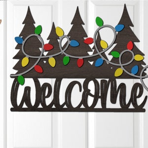 Op de afbeelding: Een houten welkomstbord met een feestelijk kerstthema. Het bord heeft een gestileerde welkomsttekst met drie dennenbomen en een reeks gekleurde lampjes.