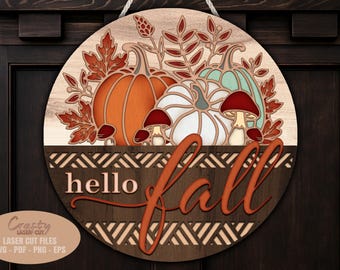 May include: Round wooden sign with a fall theme. Features an orange pumpkin, a white pumpkin, and a teal pumpkin, with red mushrooms and fall leaves. The words "hello fall" are in a cursive font. The sign is hanging on a dark brown door.