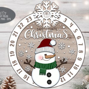 Può includere: Decorazione natalizia in legno con un pupazzo di neve con cappello rosso e sciarpa verde. Il design include un fiocco di neve e le parole "Days Until Christmas". I numeri da 3 a 23 sono visualizzati.