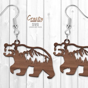 May include: A pair of wooden bear earrings with a mountain and forest silhouette design. The earrings are brown and have silver hooks.