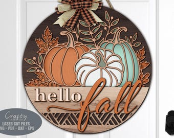 Puede incluir: Un letrero redondo de madera con las palabras "hello fall" en una fuente de escritura decorativa. El letrero está decorado con tres calabazas, dos naranjas y una azul verdosa, y hojas de otoño.