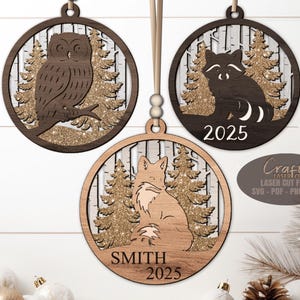 May include: Wooden Christmas ornaments with woodland animal designs. The ornaments feature an owl, raccoon, and fox, with a glittery background and tree silhouettes. The fox ornament includes the name "SMITH" and the year "2025".