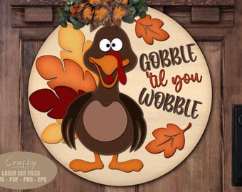Puede incluir: Un letrero de puerta redondo de madera para Acción de Gracias con un pavo de dibujos animados y el texto "GOBBLE 'til you WOBBLE". El pavo es marrón, naranja y beige, con hojas de otoño.