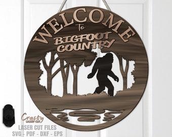 Könnte beinhalten: Ein rundes Holzschild mit dem Text "Willkommen im Bigfoot-Land" in brauner Schrift. Das Schild zeigt eine Silhouette von Bigfoot, der durch einen Wald mit Bäumen und einem kleinen Bach geht.
