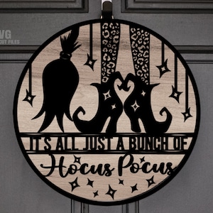 May include: A round wooden door hanger with a black silhouette of a witch's boots, a witch's hat, and stars. The text "It's all just a bunch of Hocus Pocus" is written on the door hanger.
