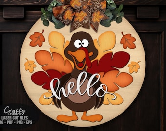 Puede incluir: Un letrero de puerta redondo de madera con un diseño de pavo en colores otoñales. El pavo tiene una carúncula roja y la palabra "hello" en escritura blanca. El letrero está decorado con hojas y un lazo.