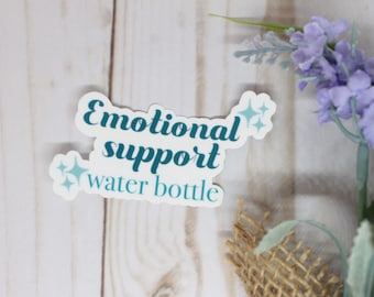Peut inclure: Un autocollant blanc avec le texte sarcelle qui dit "Emotional support water bottle". L'autocollant a deux motifs d'éclats d'étoiles sarcelle. Le fond est une surface en bois clair avec des fleurs violettes et de la toile de jute.