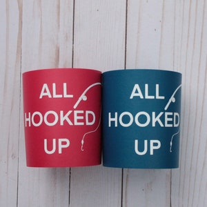 Puede incluir: Dos enfriadores de latas, uno rojo y otro azul, con el texto "All Hooked Up" y un gr&aacute;fico de ca&ntilde;a de pescar y anzuelo.