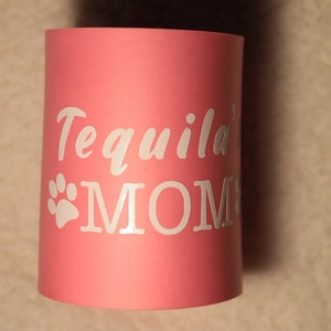 Puede incluir: Enfriador de latas rosa con texto blanco que dice "Tequila' MOM" y una huella de pata blanca.