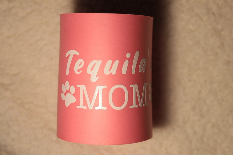 Puede incluir: Enfriador de latas rosa con texto blanco que dice "Tequila' MOM" y una huella de pata blanca.