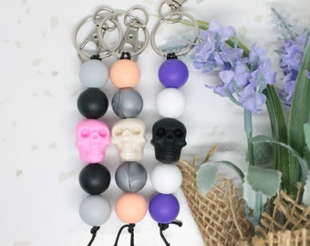 May include: Three colorful skull keychain charms with silver key rings. Each charm features a skull bead in pink, white, or black, with alternating gray, white, and peach beads.