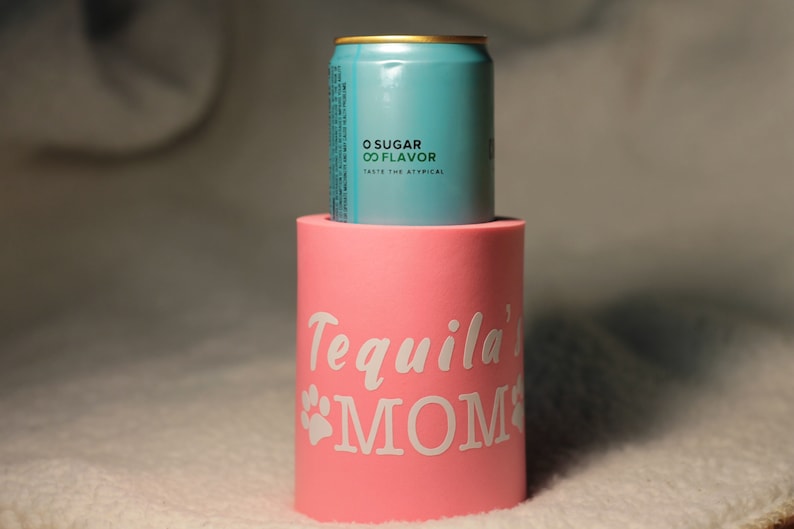 Puede incluir: Un enfriador de latas rosa con el texto "Tequila's MOM" en letras blancas y una huella de pata a cada lado. Una lata azul con el texto "0 SUGAR 00 FLAVOR TASTE THE ATYPICAL" est&aacute; sentada encima del enfriador.