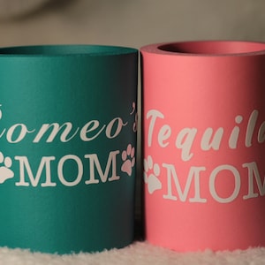 Puede incluir: Dos enfriadores de latas de espuma, uno turquesa y otro rosa, con huellas de patas blancas y el texto "Romeo's MOM" y "Tequila's MOM" respectivamente.