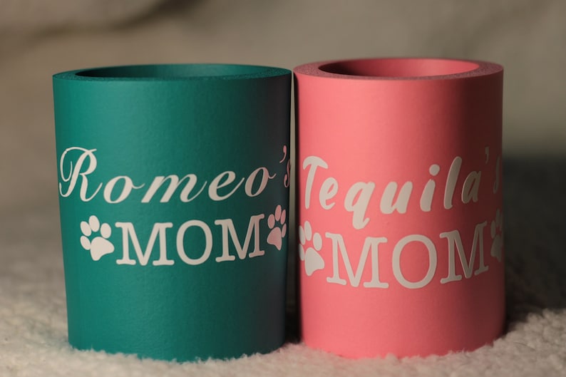 Puede incluir: Dos enfriadores de latas de espuma, uno turquesa y otro rosa, con huellas de patas blancas y el texto "Romeo's MOM" y "Tequila's MOM" respectivamente.