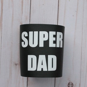 Puede incluir: Taza aislante negra y blanca con el texto "SUPER DAD" impreso en blanco.