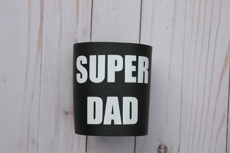 Puede incluir: Taza aislante negra y blanca con el texto "SUPER DAD" impreso en blanco.