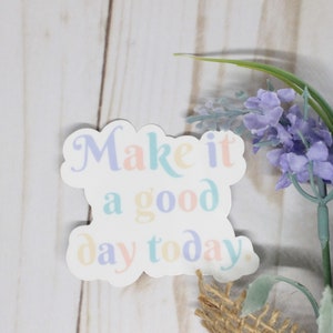 Könnte beinhalten: Ein weißer Aufkleber mit dem Text "Make it a good day today." in Pastellfarben. Der Aufkleber befindet sich auf einem weißen Holzhintergrund mit einem Zweig lila Blüten.