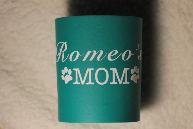 Puede incluir: Taza aislante azul turquesa con texto blanco que dice "Romeo's MOM" con dos huellas de patas blancas a cada lado de la palabra MOM.