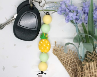 May include: A yellow pineapple charm with green leaves and white dots on a keychain with yellow, green and white beads. The keychain is attached to a set of keys and a silver key ring.