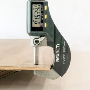 May include: A black digital caliper with a silver measuring arm is measuring a piece of wood. The caliper reads 0.15390 inches. The caliper is labeled "REXBETI" and has a measurement range of 0-25mm 0.001mm.