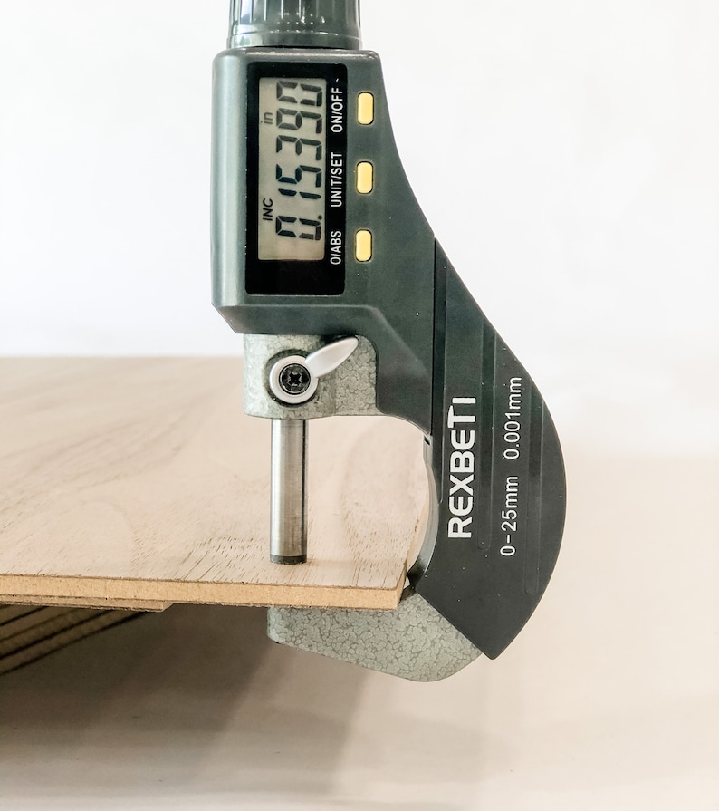 May include: A black digital caliper with a silver measuring arm is measuring a piece of wood. The caliper reads 0.15390 inches. The caliper is labeled "REXBETI" and has a measurement range of 0-25mm 0.001mm.
