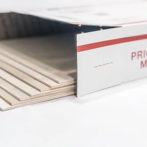 May include: A stack of thin, light brown wood panels in a white cardboard box with a red stripe and the text "PRIORITY MAIL".