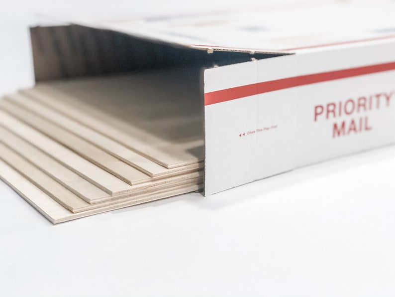 May include: A stack of thin, light brown wood panels in a white cardboard box with a red stripe and the text "PRIORITY MAIL".