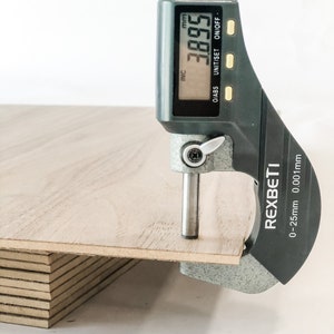May include: A close-up of a digital caliper measuring the thickness of a stack of plywood. The caliper reads 3.895 mm and is labeled REXBETI. The caliper has a range of 0-25mm with a resolution of 0.001mm.