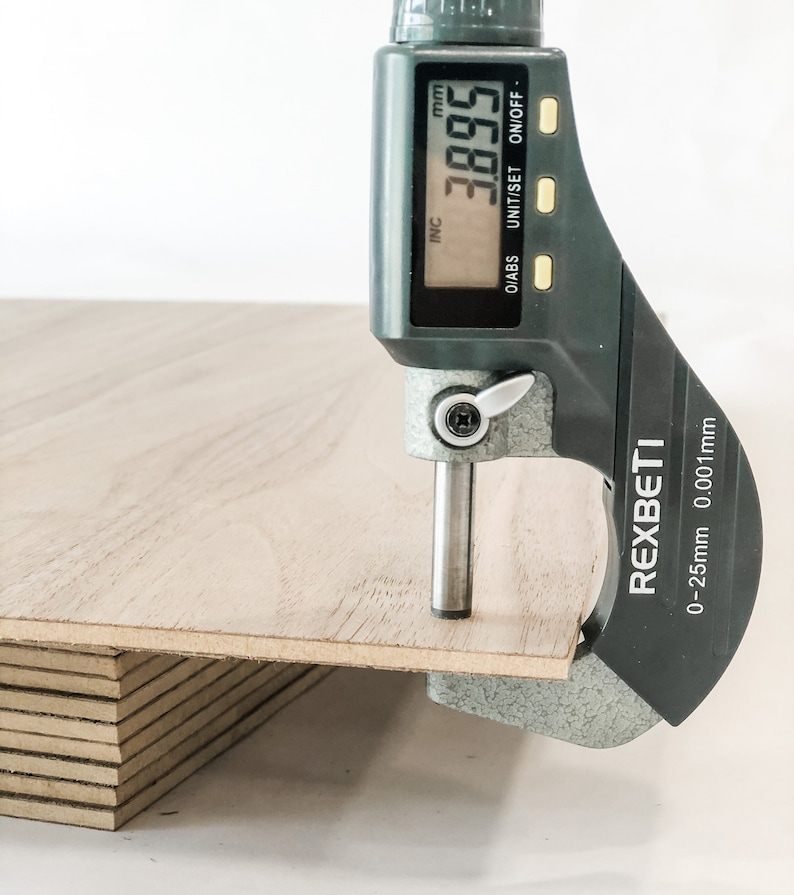 May include: A close-up of a digital caliper measuring the thickness of a stack of plywood. The caliper reads 3.895 mm and is labeled REXBETI. The caliper has a range of 0-25mm with a resolution of 0.001mm.