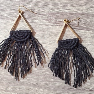 May include: A pair of earrings featuring a gold-toned triangular frame with a black macrame semi-circle attached to the base. Each earring includes a long, textured black fringe hanging from the macrame section and a gold-toned hook wire.