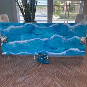 Op de afbeelding: Een rechthoekige serveerschaal met een blauw en wit harsontwerp dat oceaan golven voorstelt. De schaal heeft twee zilveren schildpadaccenten aan de uiteinden.