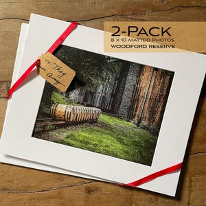May include: Two 8x10 inch matted photos of a wooden barrel in front of a brick building. The photos are packaged together with a red ribbon and a gift tag that says "To: Troy, Amy". The package is labeled "2-Pack 8 x 10 Matted Photos Woodford Reserve".