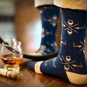 May include: Navy blue socks with a pattern of cigars and whiskey glasses. The socks have a tan heel and toe. A cigar and a glass of whiskey are on a wooden surface.