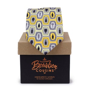 May include: A necktie with a repeating pattern of bourbon bottles and glasses in gray and black, set against a yellow and gray honeycomb design. The tie is displayed in a black box with the "Bourbon Cousins" logo and text.